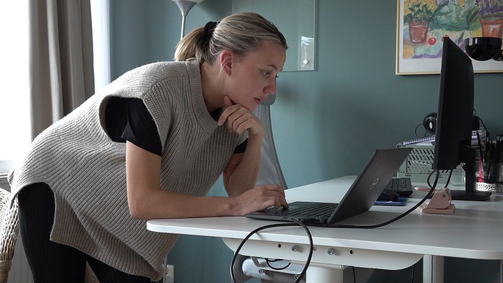 En person står lutad över en bärbar dator på ett skrivbord, med en hand på tangentbordet och den andra handen som stöd under hakan. Beskrivningen av rummet visar en lampa i bakgrunden och en vägg dekorerad med en tavla med blommor. Datorn är kopplad med sladdar och det syns även skrivmaterial på skrivbordet.