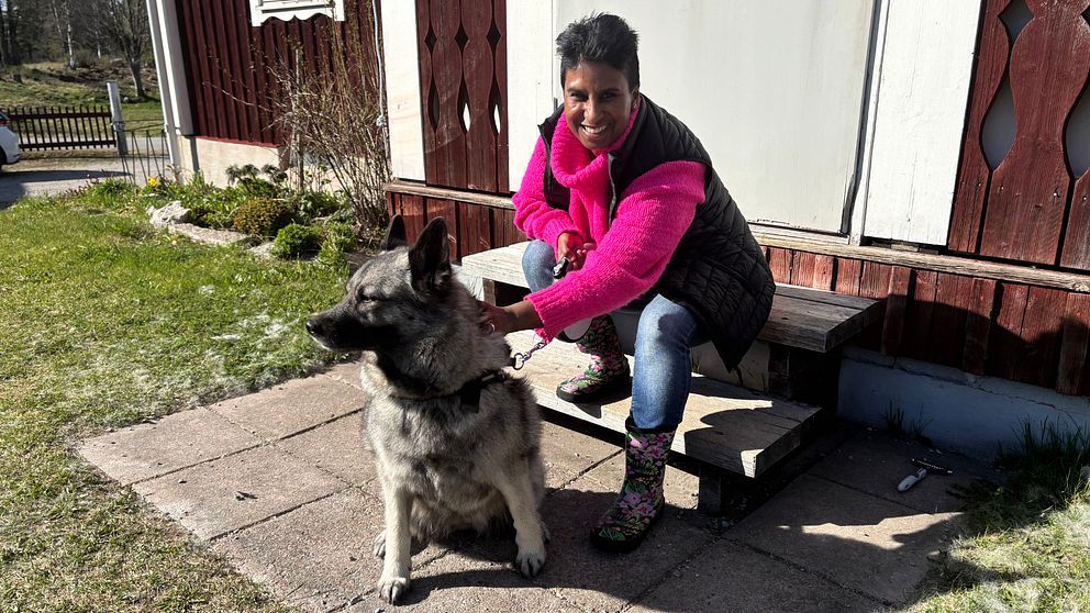 Person med rosa jacka sitter på trappa utanför hus med stor hund i koppel.