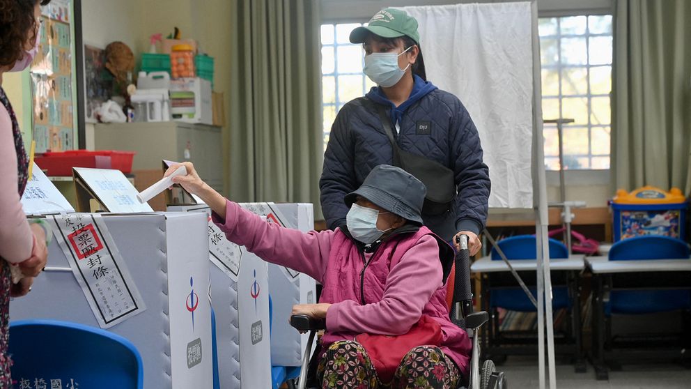 En kvinna i rullstol och blå hatt lägger sin röst i en vitt papplåda.