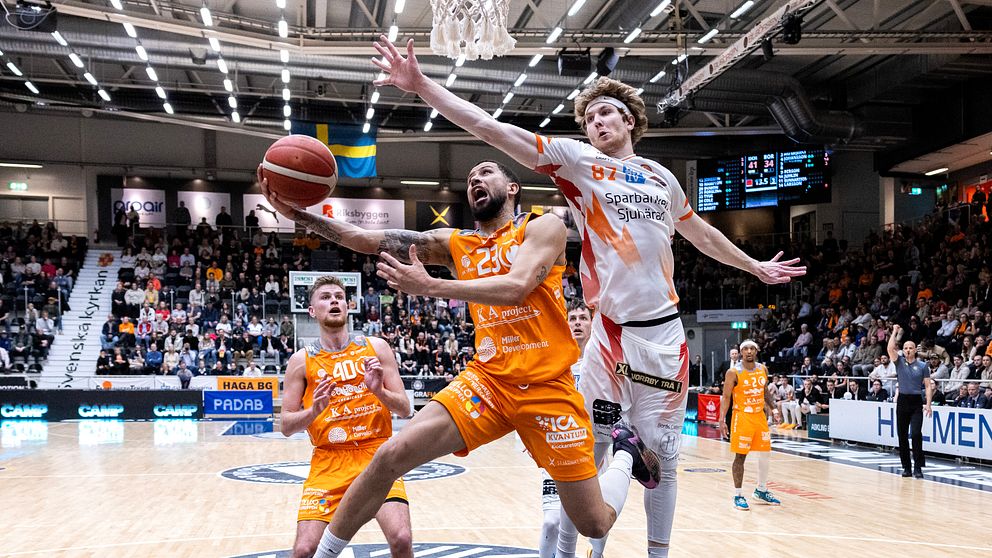 Basketspelare i orange och vitt hoppar mot korgen inför en fullsatt läktare.