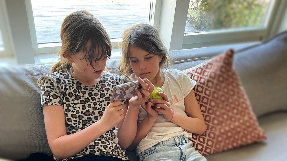 Två unga flickor sitter i soffan och tittar i en mobil tillsammans