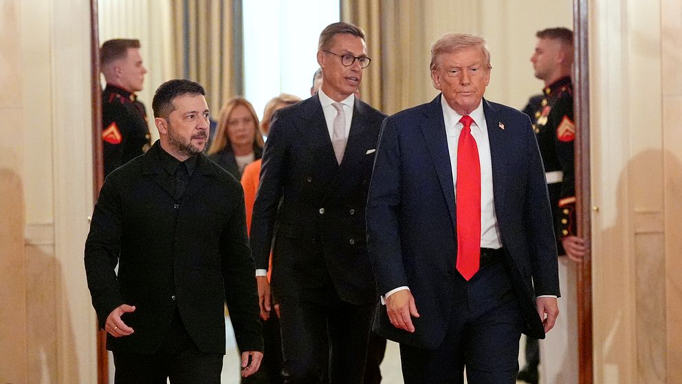 Volodymyr Zelenskyj går tillsammans med två män i kostym i en officiell byggnad.