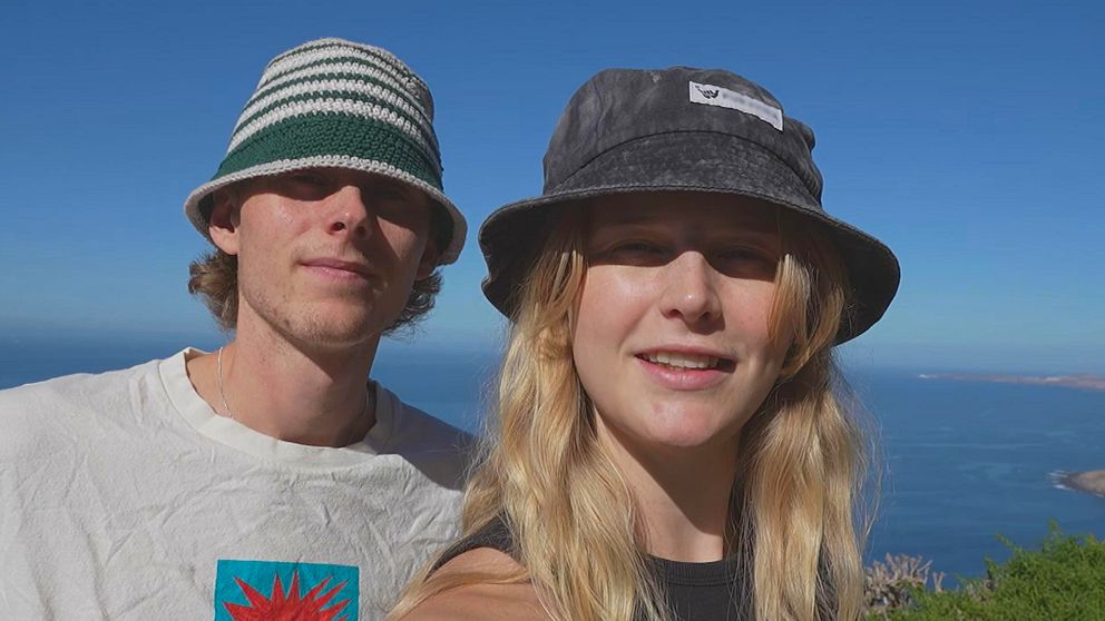 Två personer poserar framför en panoramautsikt över havet, båda med hattar och leenden. Den ena bär en vit t-shirt med färgglad design, medan den andra har på sig en svart topp. I bakgrunden syns en klar blå himmel och klippiga kustlinjer.