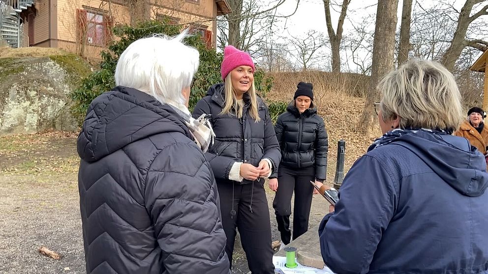 En grupp människor, varav en i rosa mössa, samtalar utomhus på en vinterdag.