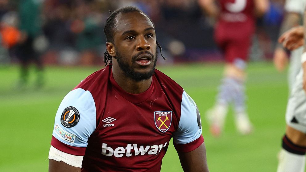 En spelare från West Ham United i en röd och ljusblå matchtröja, knäböjer på planen efter en intensiv situation under en match. I bakgrunden syns andra spelare och åskådare på stadion.