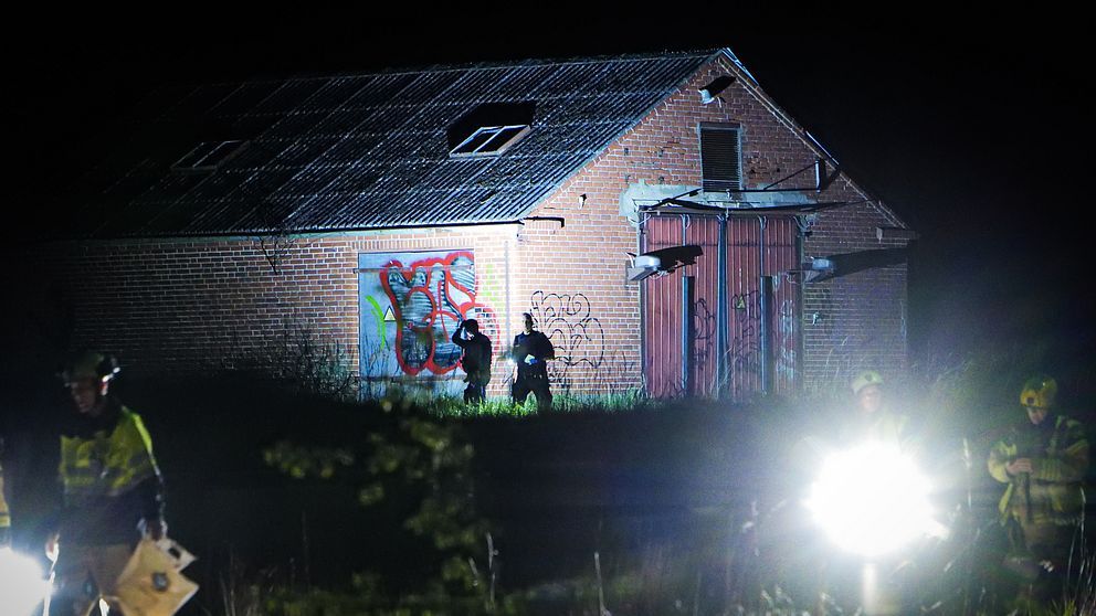 Polis undersöker området vid en nedgången lada nattetid, stark belysning i förgrunden.