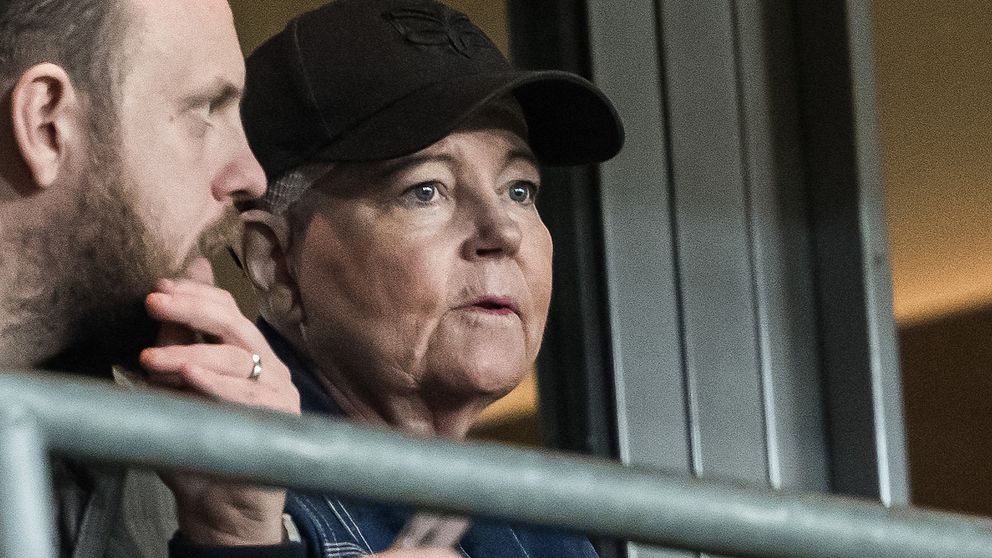 Två personer sitter i en läktare, den ena håller i en telefon och den andra ser mot en händelse i fjärran, med en stängsel i förgrunden.