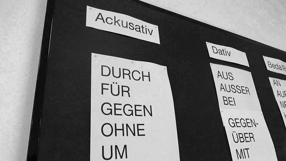 En svartvitt bild av en anslagstavla med tre rubriker: ”Ackusativ”, ”Dativ” och ”Befallning”. Under ”Ackusativ” listas prepositioner som ”DURCH”, ”FÜR”, ”GEGEN”, ”OHNE” och ”UM”. Under ”Dativ” finns prepositioner som ”AUS”, ”AUSSER”, ”BEI”, ”GEGENÜBER” och ”MIT”.