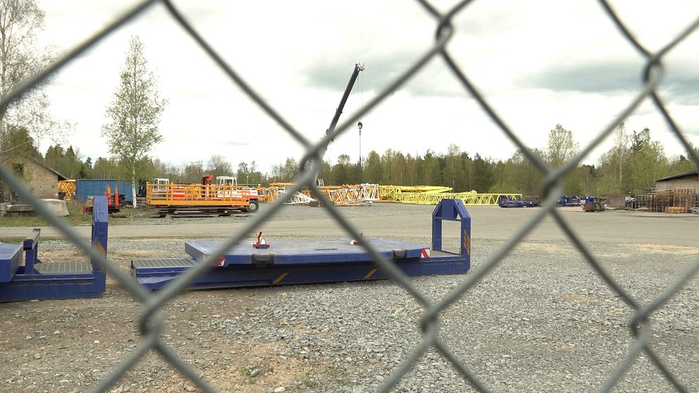Industriområde sett genom stängsel, med maskiner och fordon på grusplan.