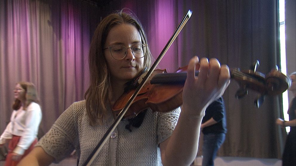 Ung person spelar fiol inomhus, omgiven av andra människor.