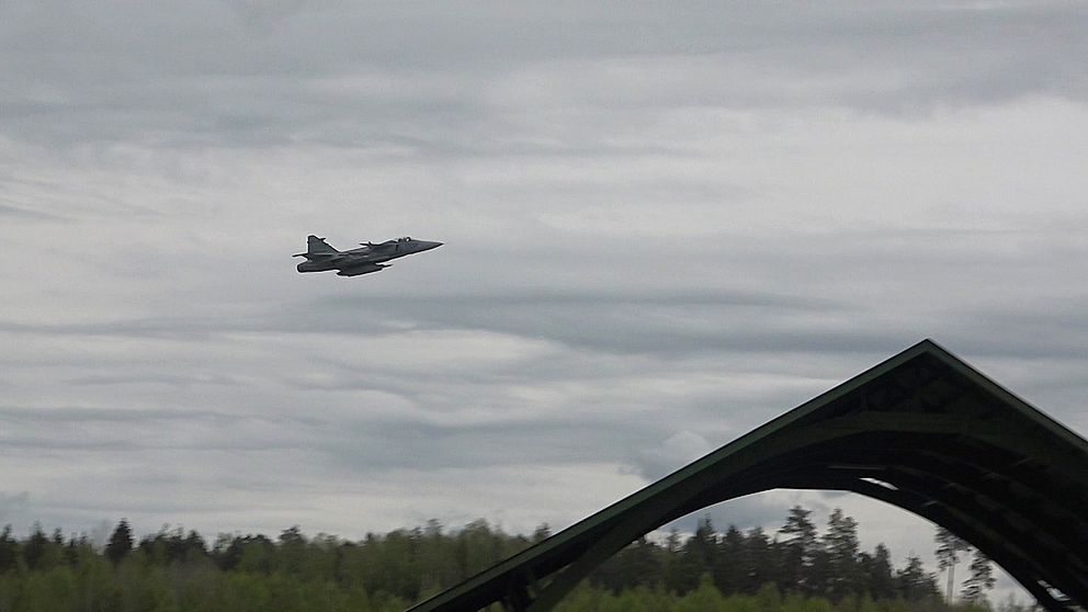 Stridsflygplan flyger lågt över skog och en militärhangar i förgrunden.