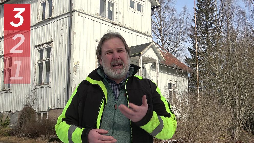 Person i gul varseljacka framför ett vitt trähus med natur i bakgrunden.