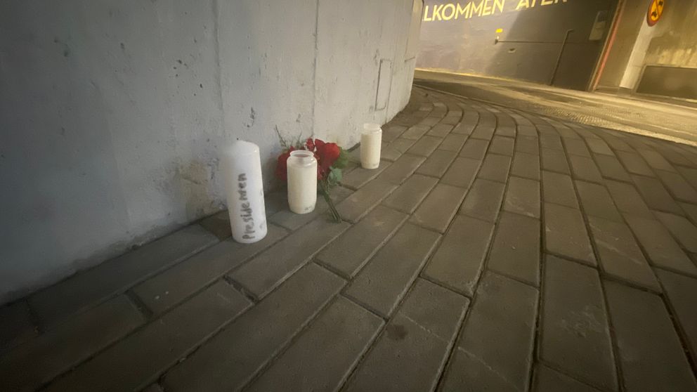 Tre ljus av olika storlekar och en röd ros ligger på en platt, gråblå stenbelagd yta. Ljusen är placerade mot en vägg, och en av dem har texten ”För alltid” skriven på. I bakgrunden syns en del av en byggnad med en ljusskylt som säger ”Välkommen åter”.