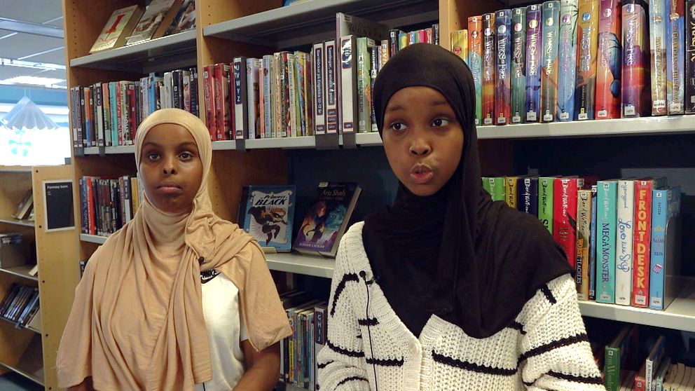 Två tjejer med hijab framför hylla med böcker i bibliotek