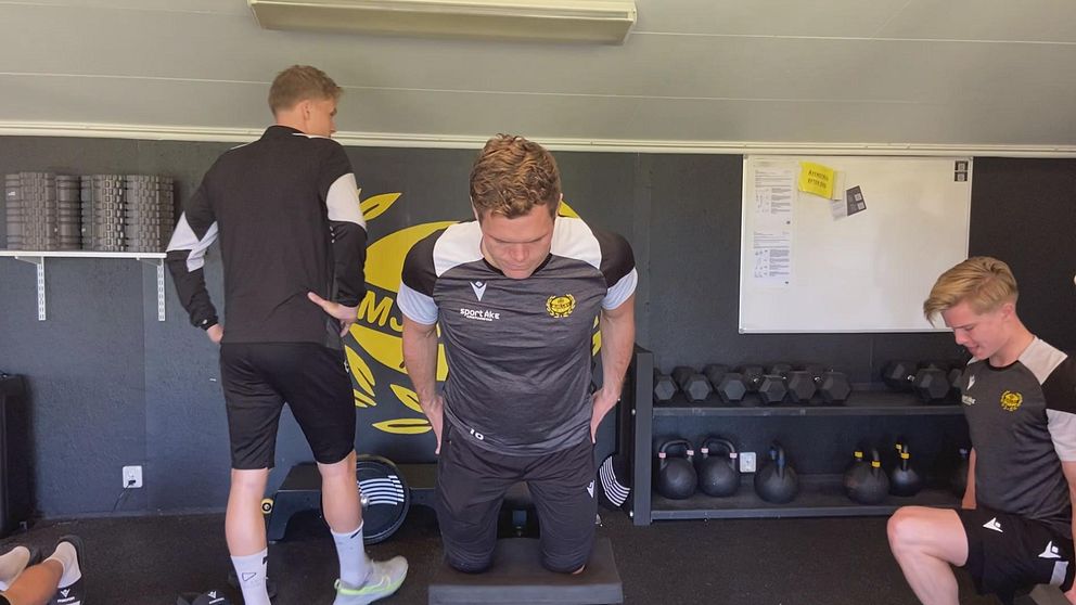Tre fotbollsspelare tränar i ett gym med kettlebells och en anslagstavla i bakgrunden.