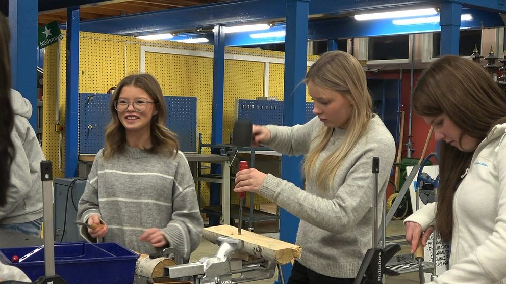 Tre personer arbetar i en verkstadsmiljö. Den första personen, som står längst till vänster, håller en penna och verkar skissa eller notera något. Den andra personen, i mitten, använder en borrmaskin på en träbit som är fastklämd i en skruvtving. Den tredje personen, till höger, håller en bit trä i ena handen och förbereder för att arbeta med den. I bakgrunden syns verktyg och en arbetsyta.