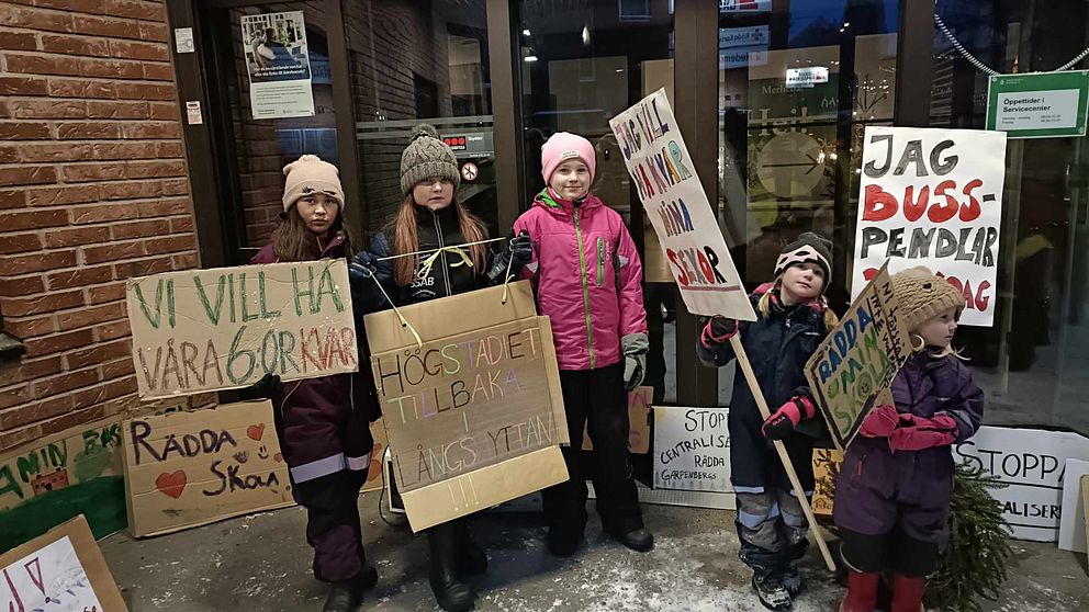 fem barn i vinterkläder, med skyltar om att bevara skolorna, framför en entré
