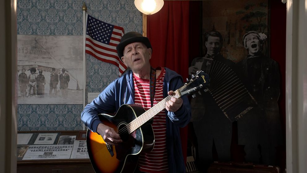 Owe ”Clapson” Carlsson spelar gitarr i ett rum som dekorerats med amerikanska flaggor, gamla fotografier och stående silhuetter av musiker med dragspel.