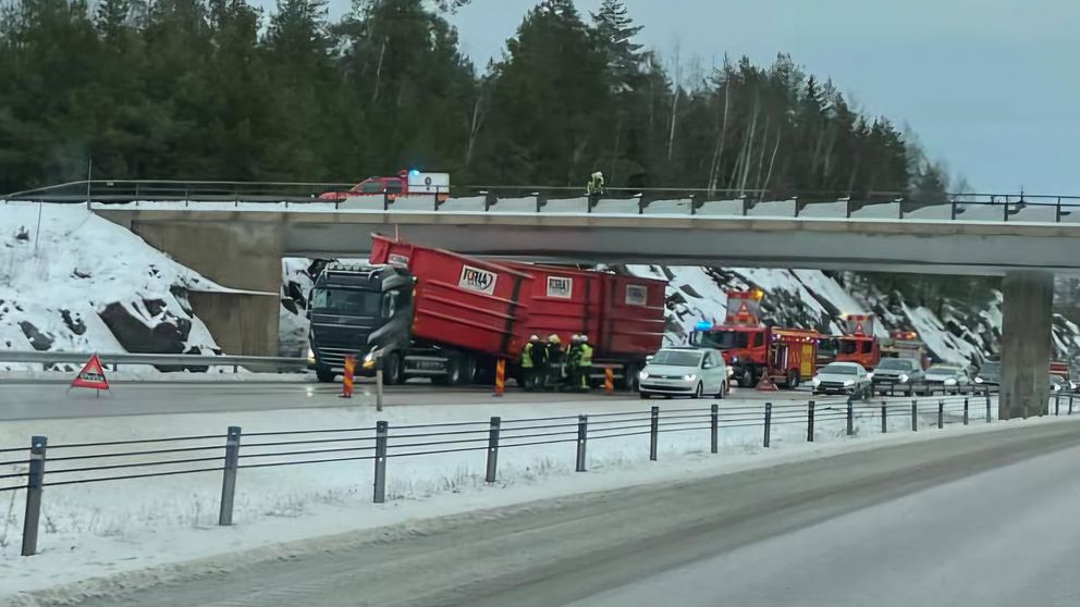 Olycka på motorväg med en lastbil som fastnat under en bro, räddningstjänst är på plats och arbetar. Flera bilar är stillastående i närheten, och snö finns i kanten av vägen.