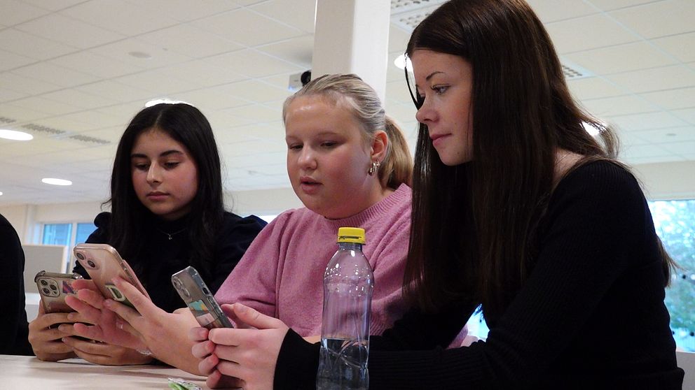 Tre personer sitter vid ett bord och tittar på sina mobiltelefoner. En av dem håller en vattenflaska. Rummet är ljust och har en modern inredning.