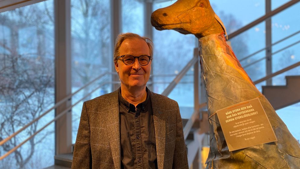 Museiechefen Olov Amelin står framför en skulptur av Storsjöodjuret på Jamtli. Utanför fönstret syns snöiga träd i bakgrunden.