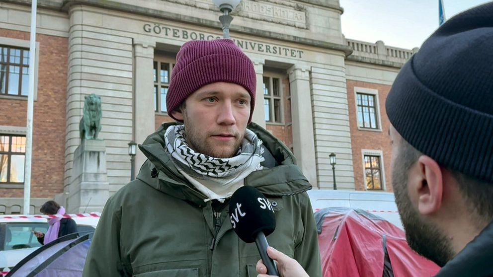 En person i en grön jacka och röd mössa intervjuas framför Göteborgs universitet. Byggnadens fasad är synlig i bakgrunden, inklusive universitetets namn. Intervjupersonen bär även en scarf över halsen och en journalist med mikrofon ställer frågor. I bakgrunden syns även tält och ett avspärrningsband.