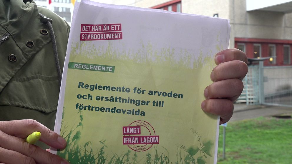 En person håller upp ett dokument med titeln ”Reglemente för arvoden och ersättningar till förtroendevalda”. Dokumentet är omslaget av grönt och gult med texten ”DET HÄR ÄR ETT STYRDOKUMENT” tydligt synlig. Bakgrunden visar en byggnad och omgivande miljö.