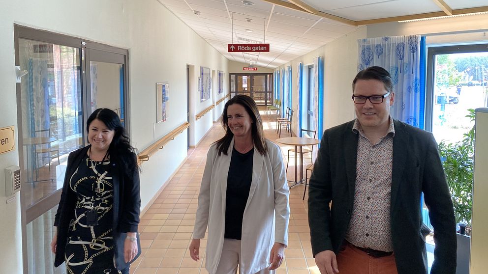 Två kvinnor går i en korridor, en klädd i svart mönstrad klänning, den andra i vit kavaj.