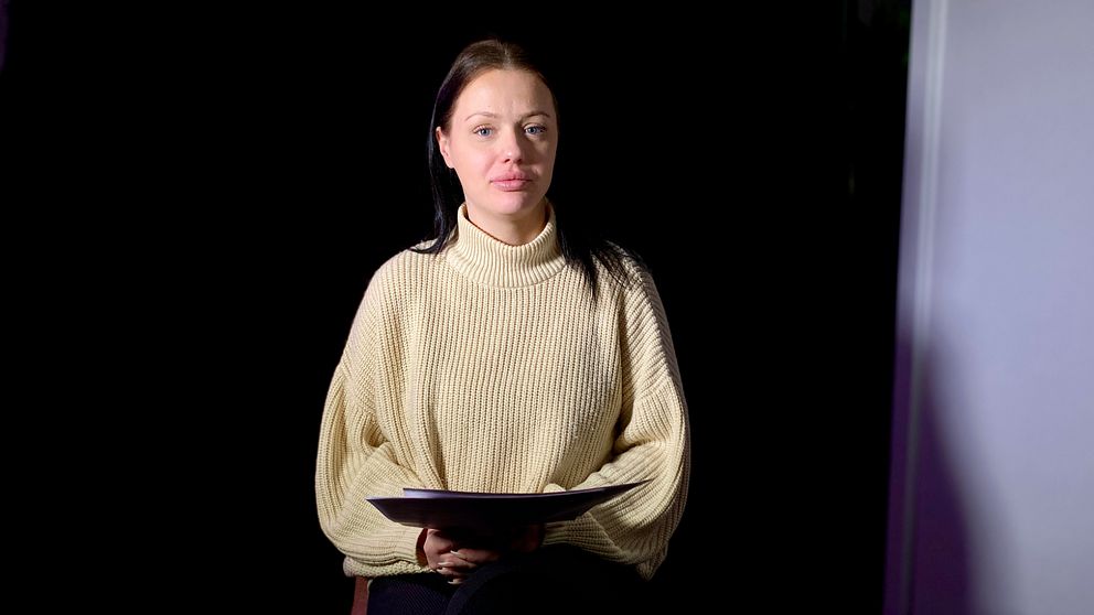 Stina sitter i en mörk miljö iförd en ljus stickad tröja och håller en som i sitt knä.