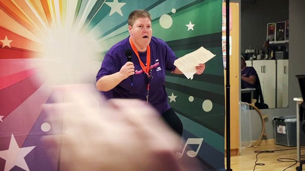 En person i lila tröja står framträdande på en scen med en färgglad bakgrund av stjärnor och musiknoter. Personen håller ett papper och gestikulerar med entusiasmerande rörelser. Framför scenen syns händer som klappar, vilket tyder på publikens uppmuntran. I bakgrunden finns en kontorsmiljö med stolar och bord.