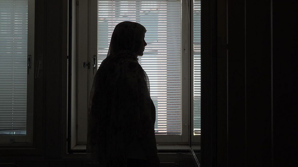 En kvinna i slöja står i profil mot ett fönster med neddragna persienner. Bilden är mörk och kvinnans ansikte syns inte