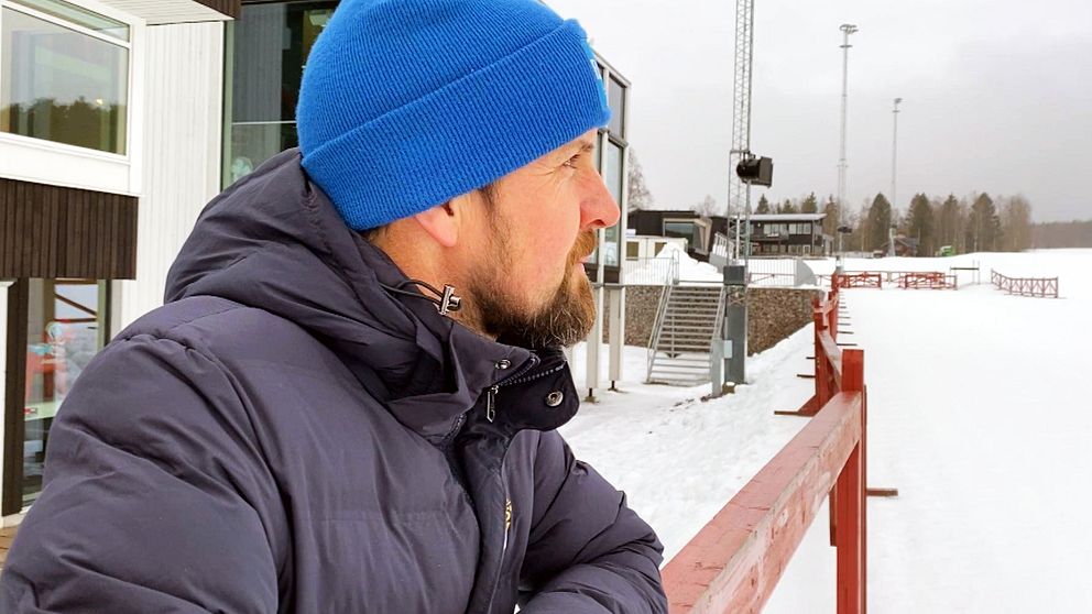 En person står utomhus iförd en blå mössa och mörk vinterjacka, med ryggen vänd mot kameran, och tittar ut över en snötäckt arena med kringliggande skog i bakgrunden.