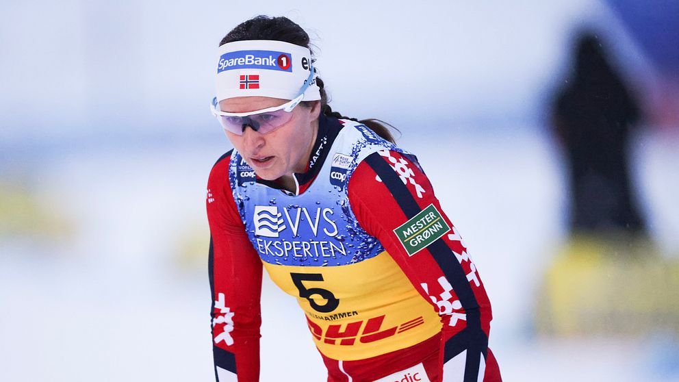 En skidåkare i röd och gul tävlingsdräkt med nummer fem, står redo på snön, förberedd för en tävling. Dräkten har sponsormärken och är utrustad för kalla förhållanden.