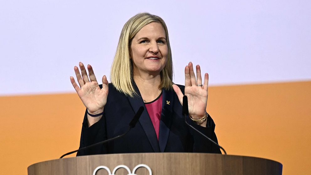 Kvinna talar vid olympisk podium med höjda händer.