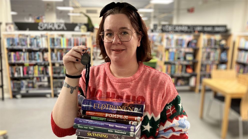 Personen håller en hög av böcker i vänster hand och en klocka i höger hand, klädd i en röd tröja med mönster. I bakgrunden syns bokhyllor i en biblioteksmiljö.