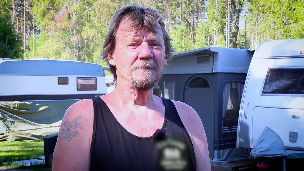 Man med linne och tatueringar står framför campingvagnar.