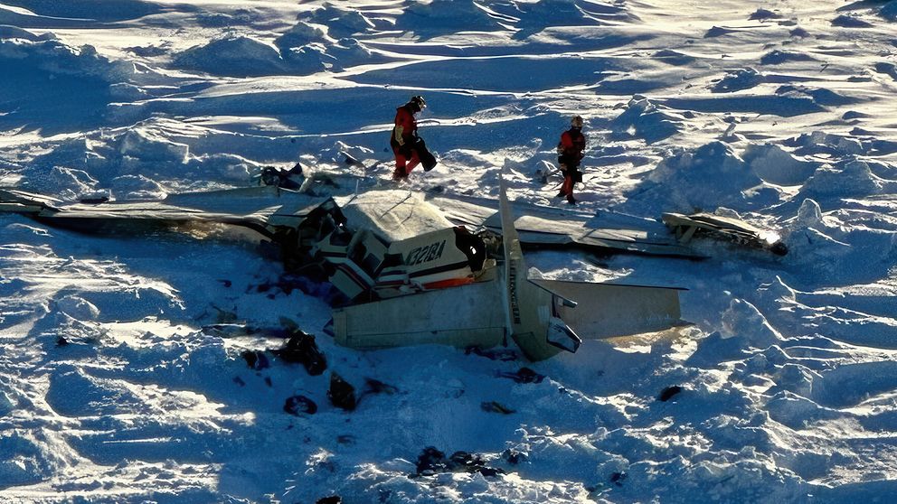Två personer i räddningskläder undersöker vraket av ett litet flygplan som har kraschlandat på ett snötäckt fält.