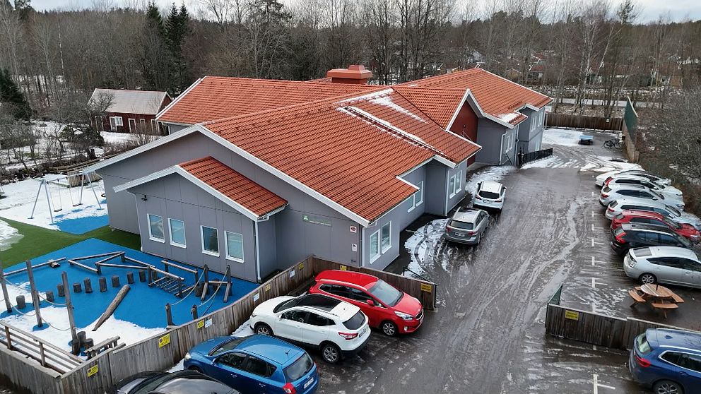 Vy över en skolbyggnad med röd takpannor, omgiven av en lekplats och en parkeringsplats med flera bilar. Lekplatsen har en blå yta och gungor, och i bakgrunden syns en skog med träd.