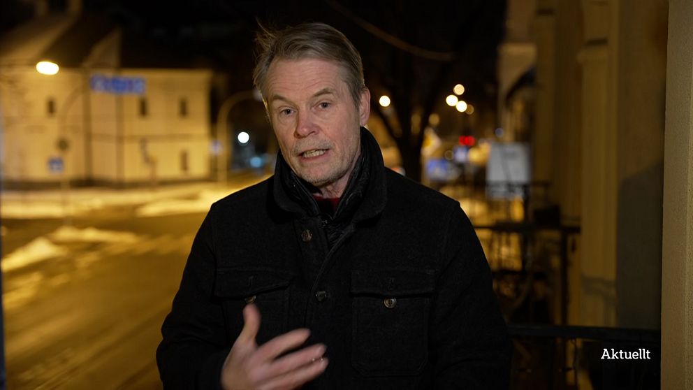 Person klädd i mörk jacka gestikulerar på en gata i en stad på kvällen.