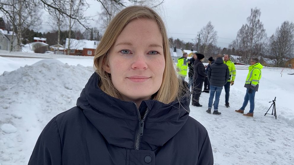 En vintermiljö med snö på marken. I bakgrunden samlas flera personer, några i neongula jackor, förmodligen del av ett arbetslag eller en grupp vid ett utomhusevenemang, framför en större snödriva omgiven av träd och byggnader.