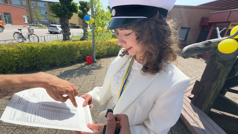 Student i vit kostym och mössa får dokument visat för sig utomhus på skolavslutning.