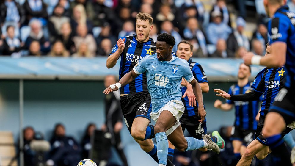 En fotbollsspelare i ljusblå tröja springer med bollen, jagad av motståndare i blåsvarta dräkter.