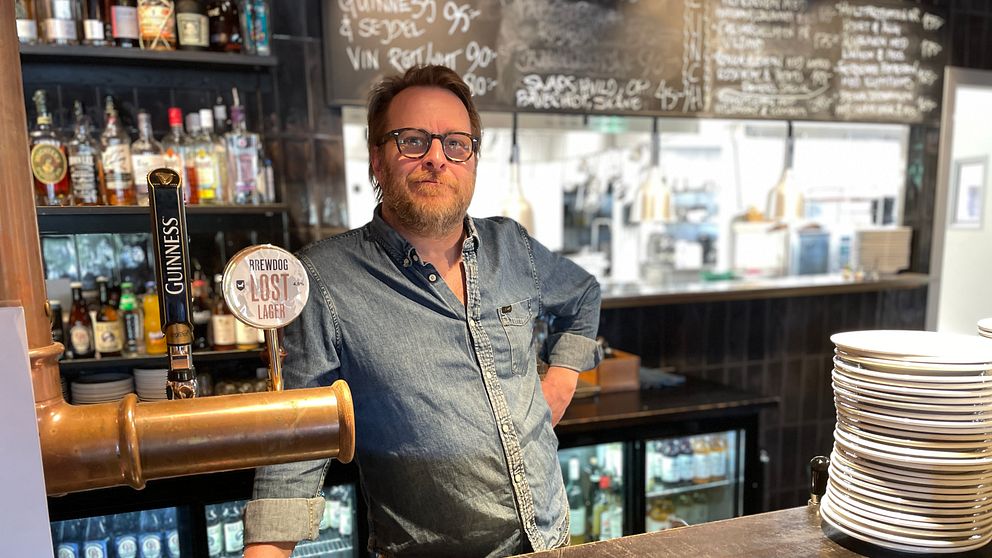 Bartender står bakom en bardisk med flaskor, ett ölkran och staplade tallrikar.