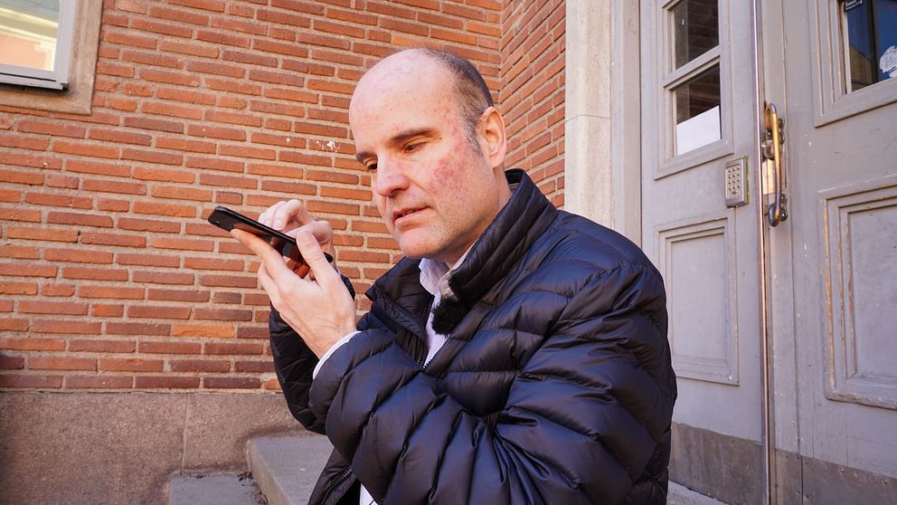 Halvbild på Fredrik Larsson som sitter på en stentrappa. Han håller upp en mobil nära ansiktet, har mörkt hår och en svart jacka. I bakgrunden ett tegelhus och en dörr.