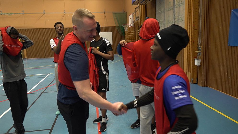 En grupp ungdomar i sportkläder befinner sig i en inomhusidrottshall. Politikerna Per Fredriksson skakar hand med en kille.