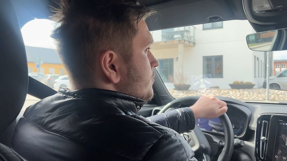 En person kör bil, hand på ratten. Byggnader syns utanför bilens fönster.