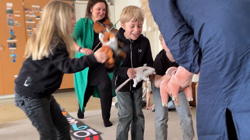 Barn leker med mjukisdjur medan en vuxen spelar fiol i bakgrunden.