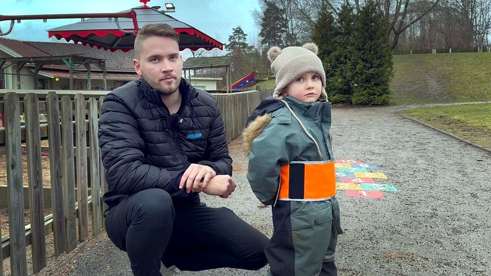 En vuxen och ett barn står vid en lekplats utomhus. Barnet bär en grön overall med en orange reflexväst och en mössa med öron. Lekplatsen har en färgglad lekställning i bakgrunden och omgivningen är grön och skuggigare med träd och gräsmatta.