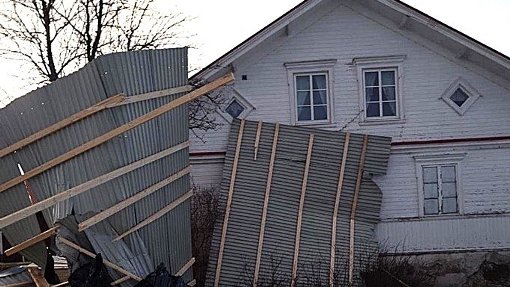 Ett stort plåttak har blåst mot ett gammalt vit timmerhus.  Bakom huset syns en trädtopp och en klar himmel, vilket tyder på att det är en solig dag.