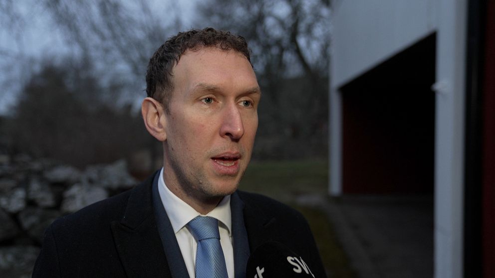 En man i kostym står utomhus i en dyster miljö med träd och en byggnad i bakgrunden. En mikrofon märkt ”SVT” syns framför.